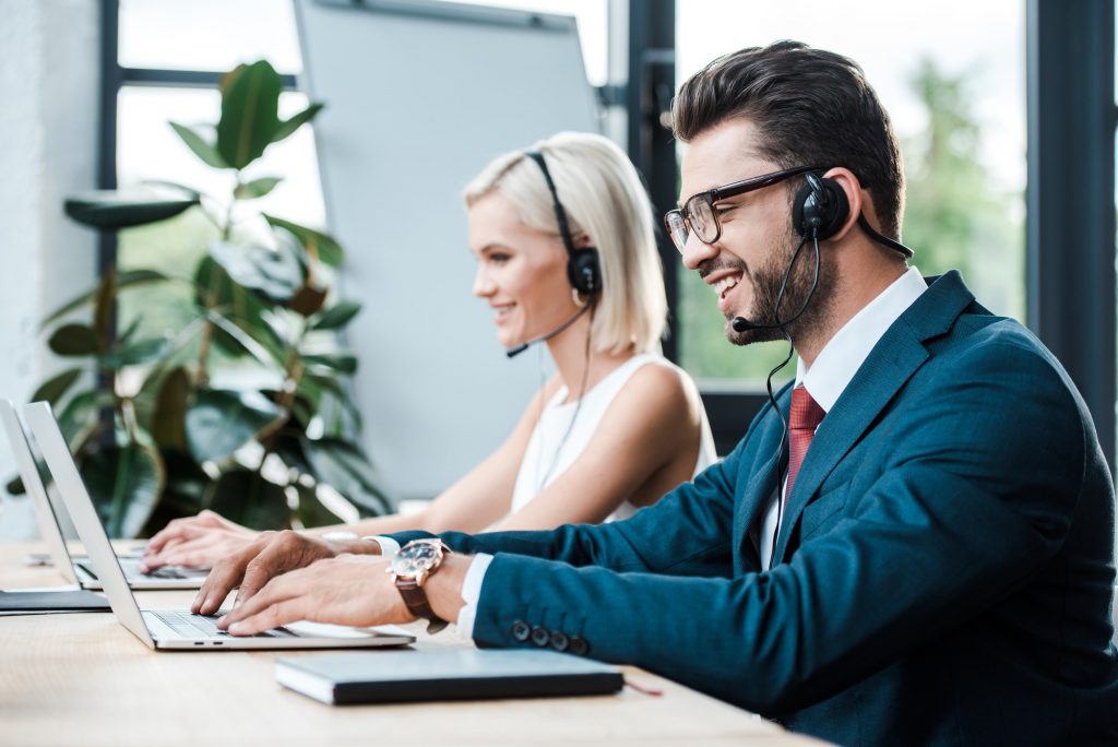 selective focus of happy operator in headset typing on laptop near blonde colleague in headset